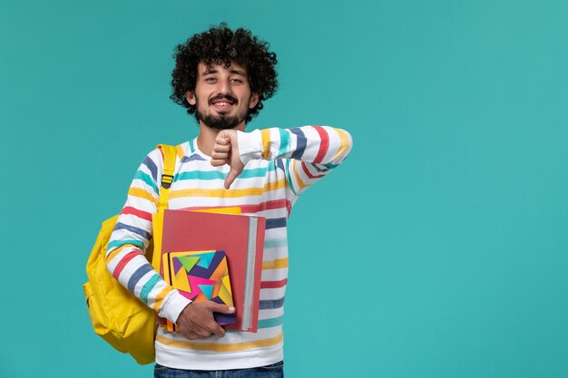 Front view of male student wearing yellow backpack holding files and copybooks on blue wall