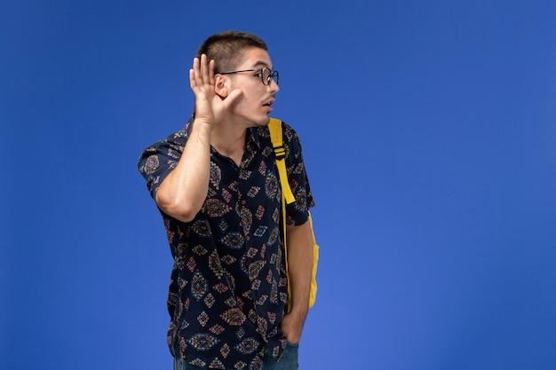 Front view of male student wearing yellow backpack trying to hear on the blue wall