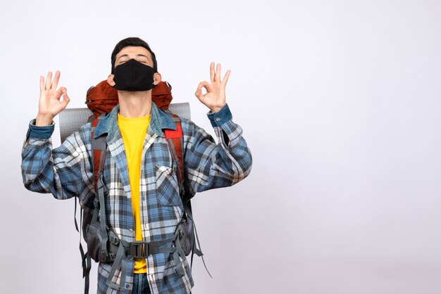 Front view male traveler with backpack and mask making ok sign with closed eyes