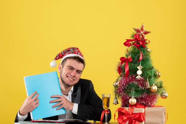 Front view male worker holding documents around little xmas tree and presents on a yellow 