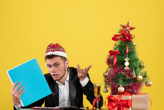 Front view male worker holding documents around little xmas tree and presents on a yellow 