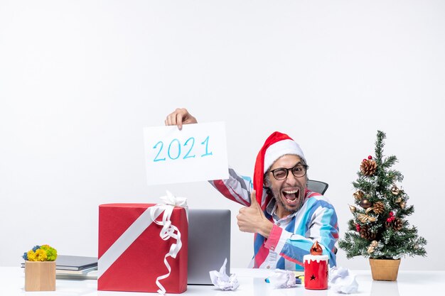 Front view male worker sitting in his working place holding paper sheet with number 2021, new year concept