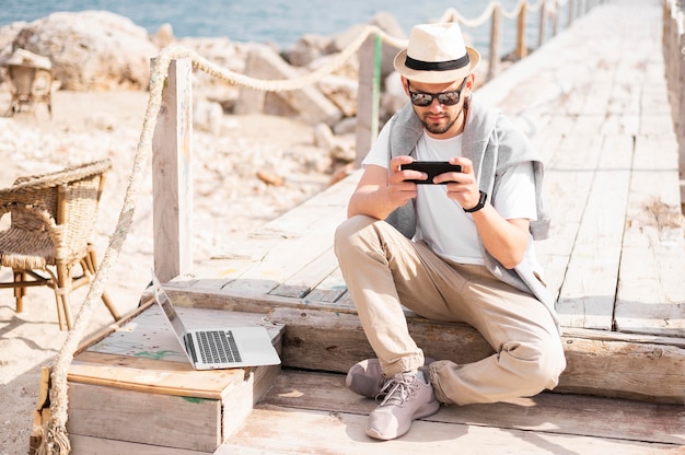 Front view of man on beach pier working on smartphone with laptop