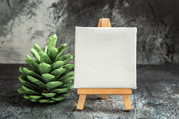 Free photo front view mini white canvas with wooden easel green pinecone on dark