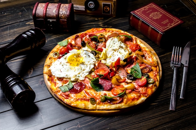 Free photo front view mixed pizza with tomatoes olives bell pepper eggs sausages on the board a book a knife and a fork on the table