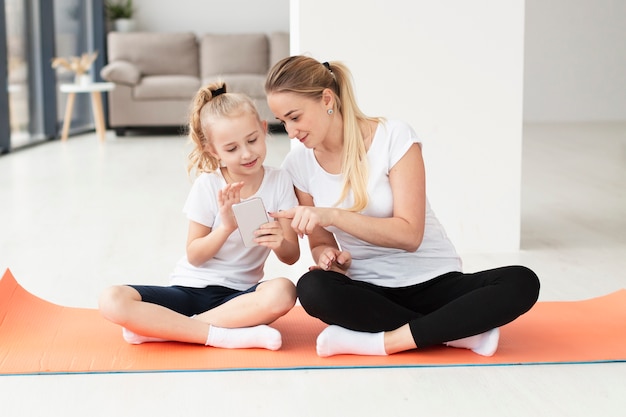 Front view of mother and daughter at home on yoga mat playing on smartphone