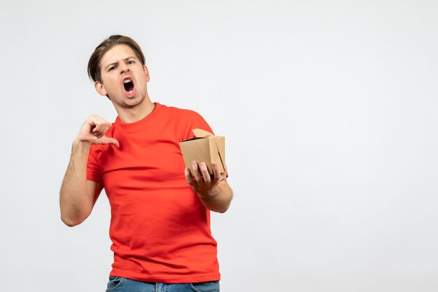 Front view of nervous young guy in red blouse holding small box pointing himself on white background