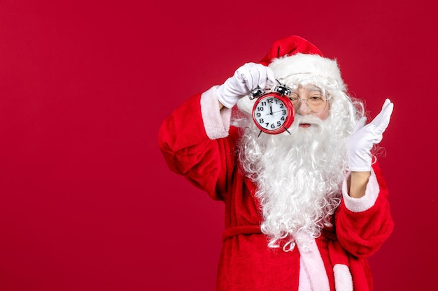 Free photo front view of old santa claus in red suit holding clock on red wall