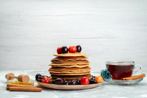 A front view pancakes with cherries inside plate with cinnamon and tea on the dark background fruit cake biscuit bake