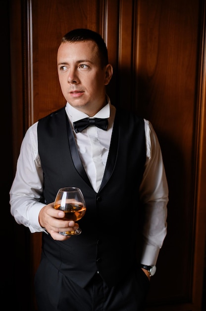 Free photo front view of pensive bridegroom wearing in stylish black suit and white shirt with bow tie holding glass filled with drink