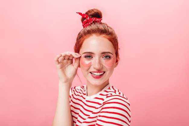 Free photo front view of playful ginger girl with eye patches. studio shot of young woman doing skincare treatment with smile.