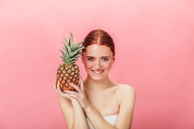 Front view of pleasant girl with exotic fruit. Studio shot of ginger young woman with pineapple isolated on pink background.