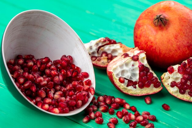 Front view of pomegranate berries spilling out of bowl and whole pomegranate with pieces around on green surface