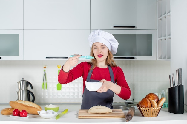 Front view pretty female chef in cook hat and apron whisking egg in the kitchen