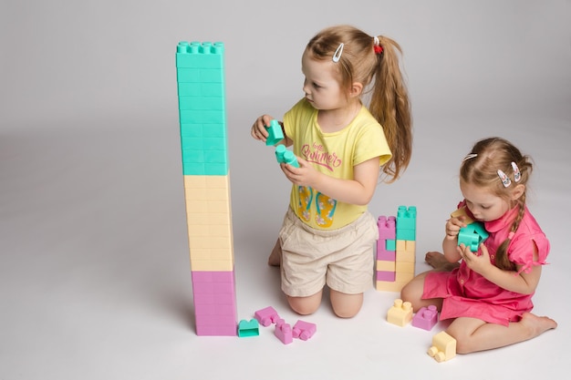 Free photo front view of pretty sisters keeping hands together looking at camera and smiling on isolated background lovely girls playing with building toys and enjoying game concept of happiness and joy