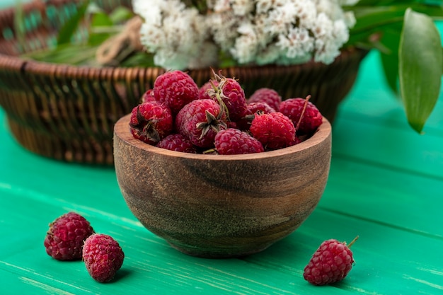 Front view of raspberries in a wooden bowl with flowers on a green surface