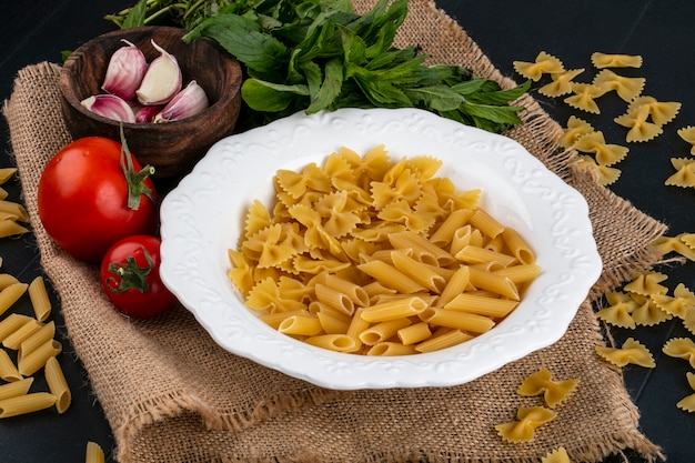 Front view of raw pasta in a plate with tomatoes garlic and a bunch of mint on a beige napkin