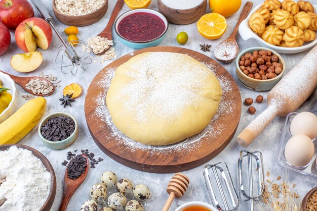 Front view of raw pastry flour on round board grater fresh fruits jam cookies hazelnuts on stained white background