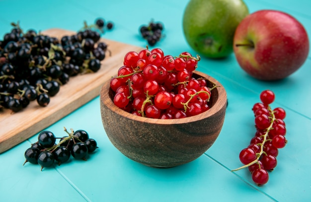 Front view red currants in a bowl with black currants on a board and apples on a light blue background