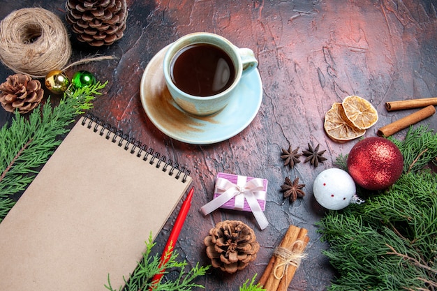 Front view red pen a notepad pine tree branches xmas tree ball toys and gift cup of tea cinnamon anises straw thread on dark red background