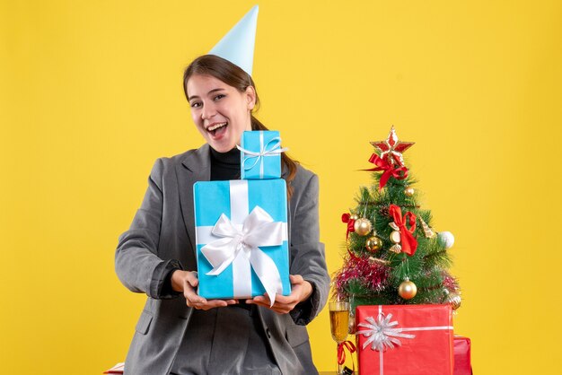 Free photo front view rejoiced girl with party cap holding xmas gifts with both hands near xmas tree and gifts cocktail