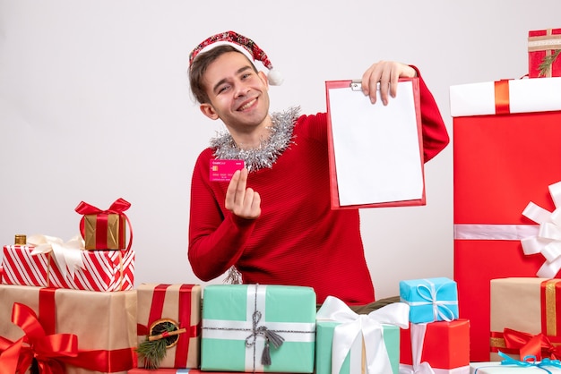 Free photo front view rejoiced young man holding clipboard and card sitting around xmas gifts