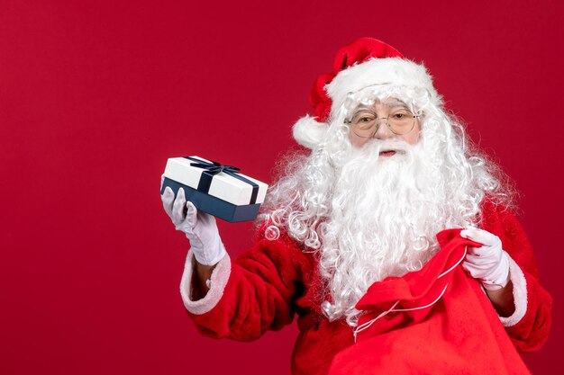 Front view santa claus holding present from bag full of presents for kids on red floor new year