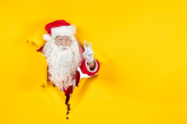 Free Photo front view of santa claus looking through ripped paper yellow wall