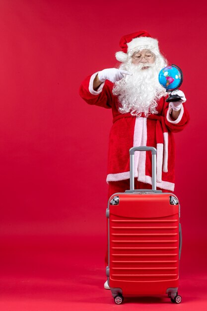 Front view of santa claus with bag holding little globe and preparing for trip on red wall