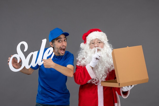 Free Photo front view of santa claus with male courier holding sale banner and food box on grey wall