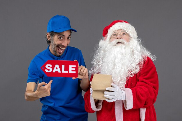 Front view of santa claus with male courier holding sale banner and food packages on a grey wall