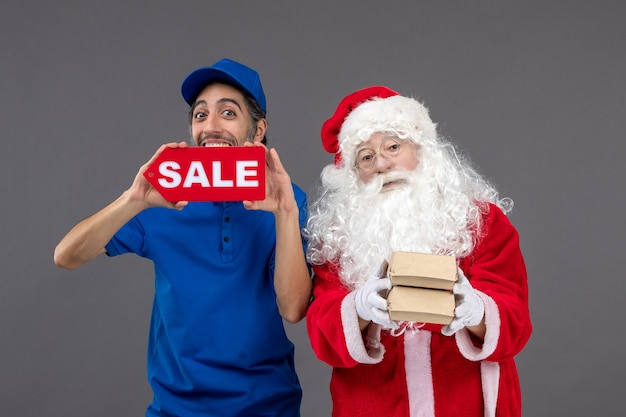 Front view of santa claus with male courier holding sale banner and food packages on grey wall