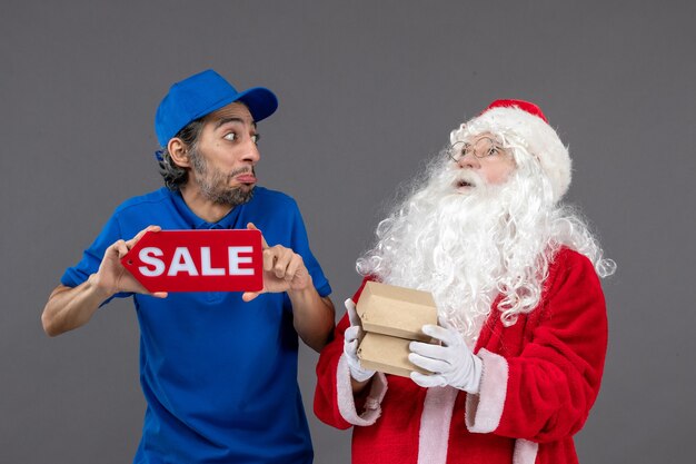 Front view of santa claus with male courier holding sale banner and food packages on grey wall