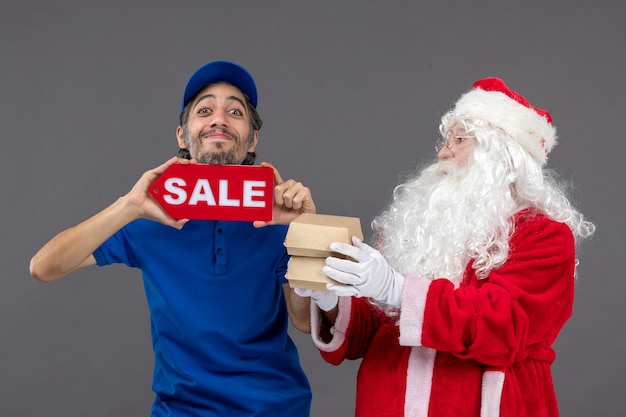 Free Photo front view of santa claus with male courier holding sale banner and food packages on grey wall