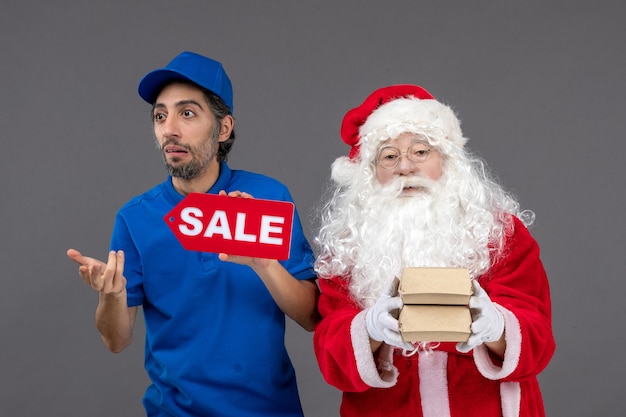 Front view of santa claus with male courier holding sale banner and food packages on grey wall