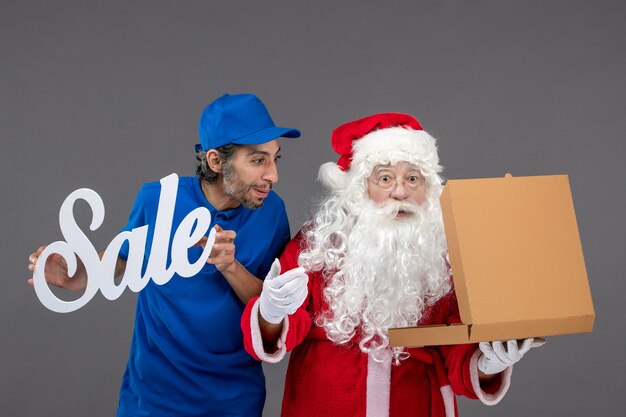 Front view of santa claus with male courier holding sale banner and opening food box on a grey wall