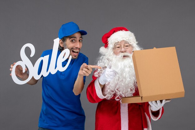 Front view of santa claus with male courier holding sale banner and opening food box on grey wall