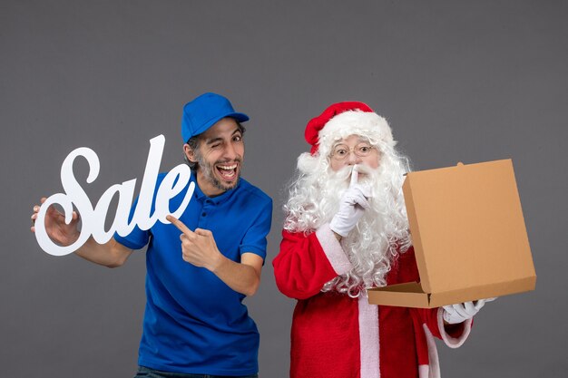 Front view of santa claus with male courier holding sale writing and food box on grey wall