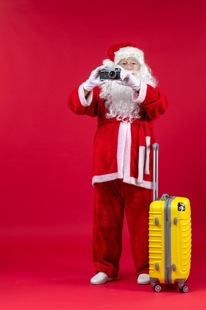 Front view of santa claus with yellow bag taking photo with camera on red wall