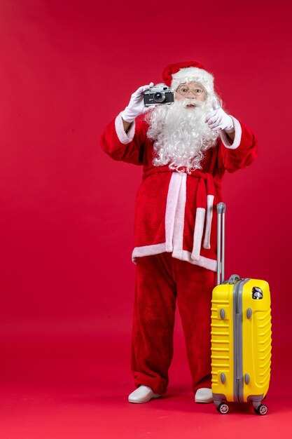 Front view of santa claus with yellow bag taking photo with camera on red wall