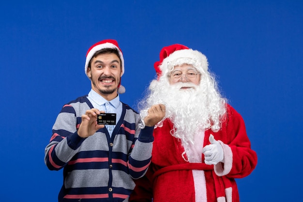 Front view of santa claus with young man holding bank card on blue wall