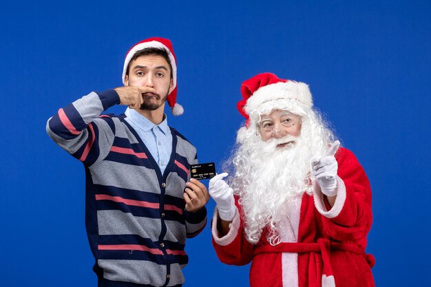 Front view of santa claus with young man holding bank card on blue wall