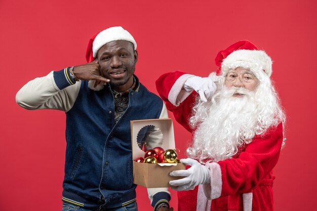 Front view of santa claus with young man holding xmas tree toys on a red wall