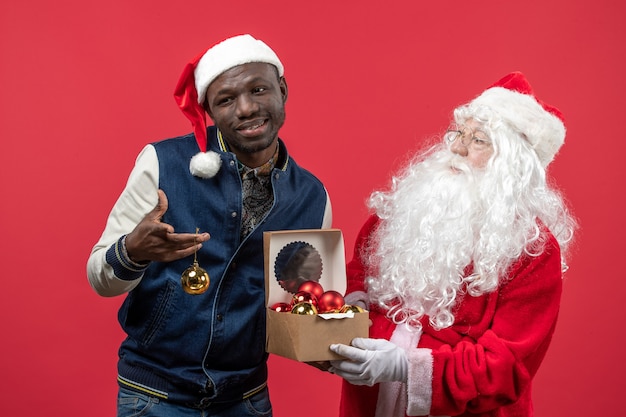 Front view of santa claus with young man holding xmas tree toys on a red wall