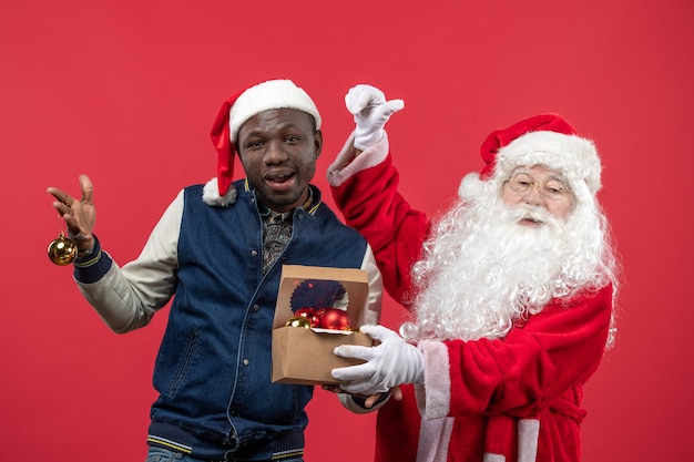 Free photo front view of santa claus with young man holding xmas tree toys on red wall