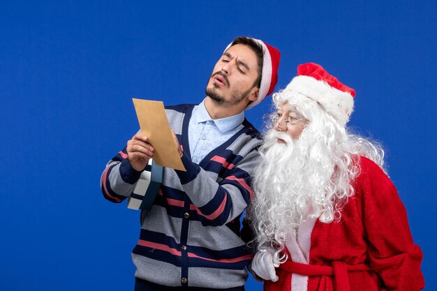 Front view of santa claus with young man whos reading letter on blue wall