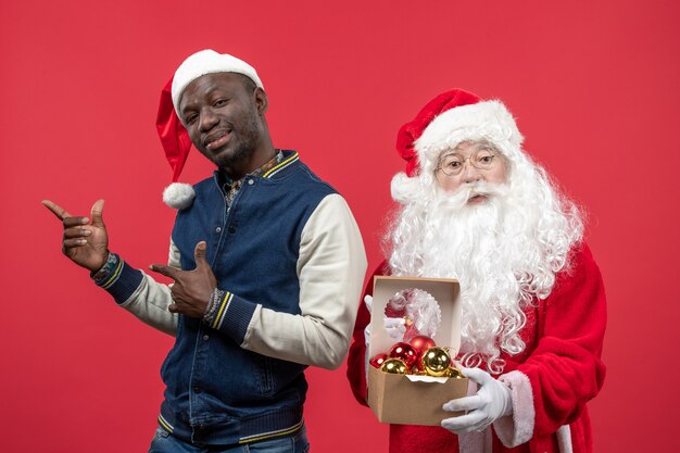 Front view of santa claus with young young man holding xmas tree toys on red wall