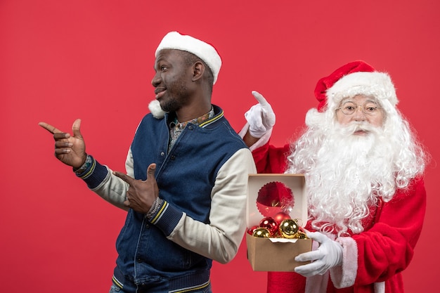 Front view of santa claus with young young man holding xmas tree toys on the red wall