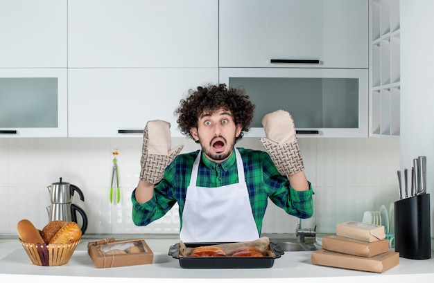 Free Photo front view of scared guy wearing holder standing behind table with freshly-baked pastry on it in the white kitchen