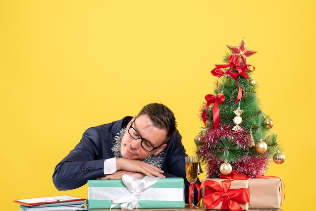 Front view of sleepy business man sitting at the table near xmas tree and presents on yellow